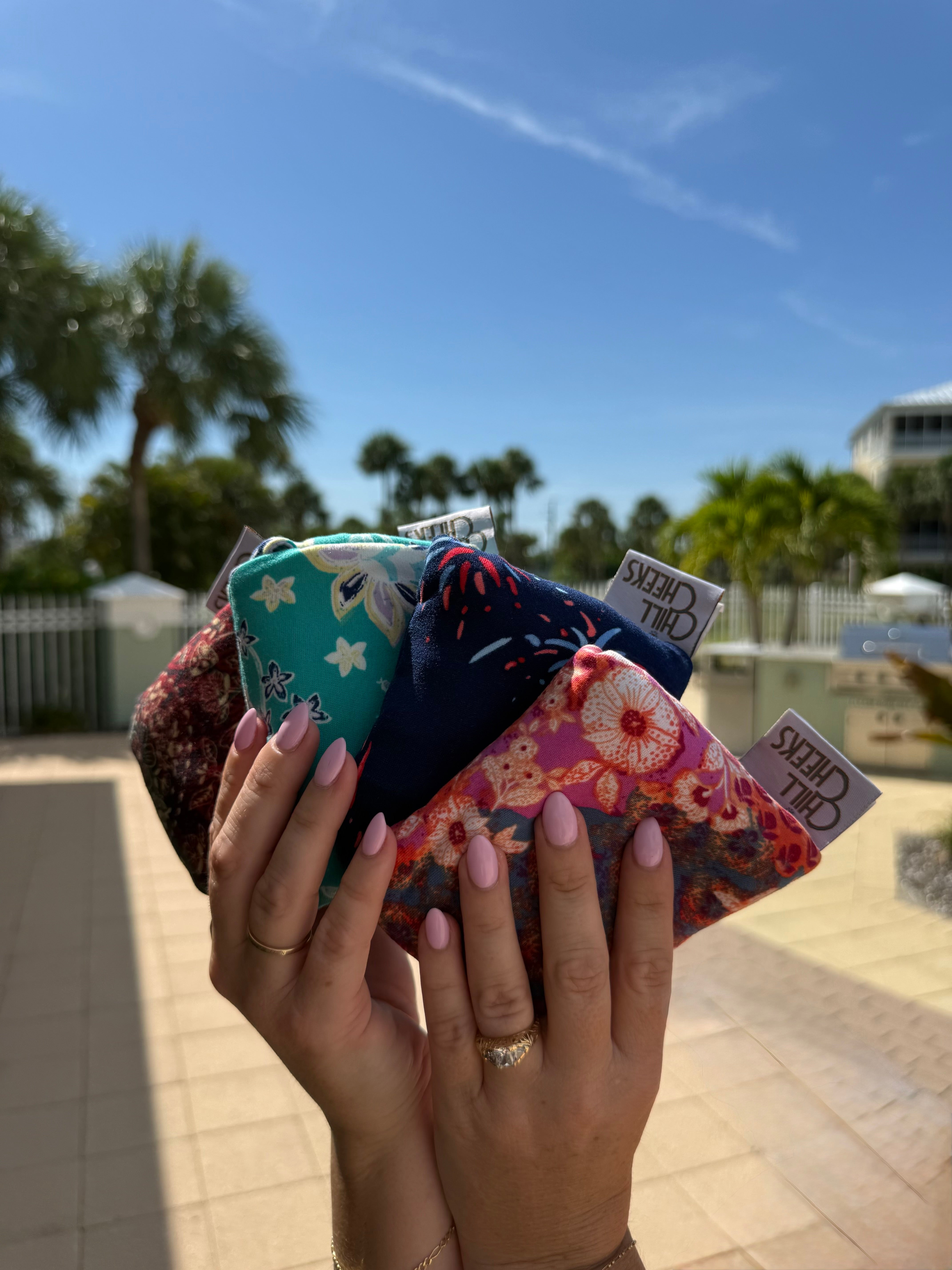 A person holds a cluster of Chill Cheeks, folded into the integrated fabric pouch, in multiple floral and patterned prints. Photographed outdoors with palm trees, a blue sky, and a patio in the background. The pouches show visible 'Chill Cheeks' tags.
